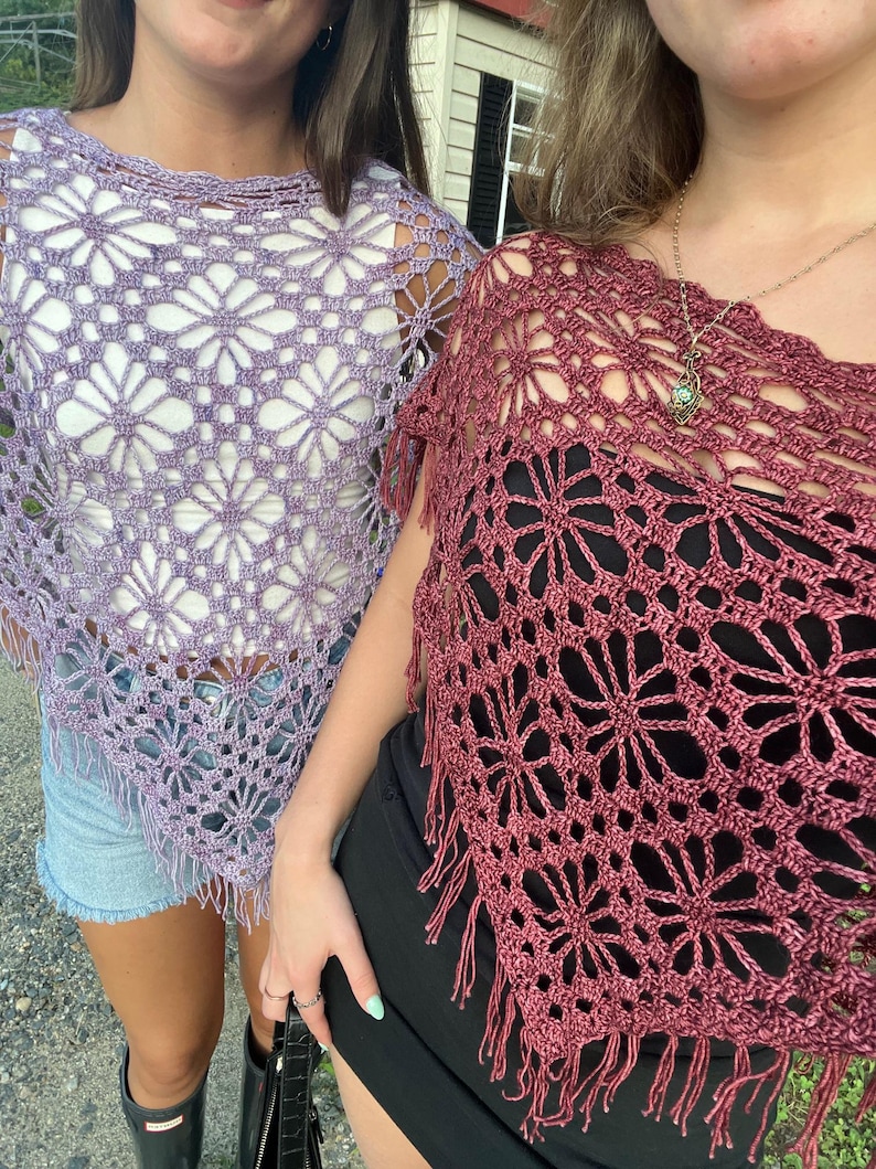 May include: Two crochet ponchos are shown. One is a light lilac colour, and the other is a deep red-purple. Both have a floral pattern and fringe along the bottom. The ponchos are worn over other clothing.