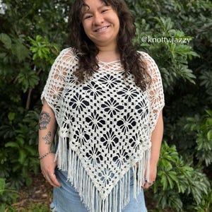 May include: Cream crochet poncho featuring a floral design and fringe trim. The openwork pattern reveals a dark top beneath. The poncho is worn with denim shorts. The text "@knotty.jazzy" is visible.