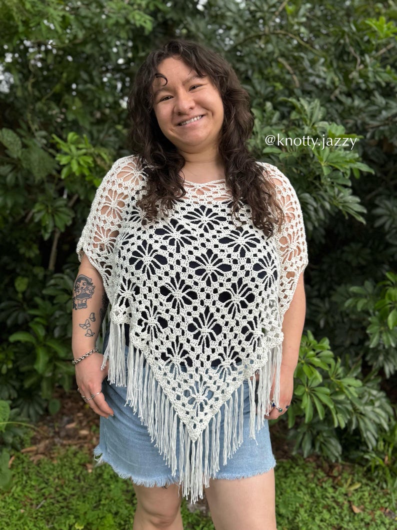 May include: Cream crochet poncho featuring a floral design and fringe trim. The openwork pattern reveals a dark top beneath. The poncho is worn with denim shorts. The text "@knotty.jazzy" is visible.
