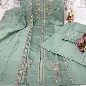 May include: A sage green three-piece outfit consisting of a long-sleeved tunic with a V-neckline, matching trousers, and a dupatta. The tunic features intricate floral embroidery in pink, yellow, and blue. Text on the image reads S TO 3XL Nayra.