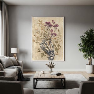 May include: A framed art print featuring a skeletal hand holding a bouquet of flowers. The flowers are a mix of purple and yellow, set against a beige background. The artwork has a vintage, gothic aesthetic, suitable for home decor.