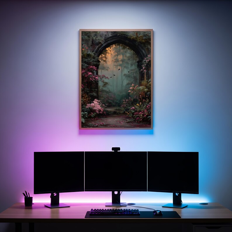 May include: A framed artwork depicting a mystical archway in a lush garden, with pink and green foliage. The artwork hangs above a desk with three black computer monitors, a keyboard, and a mouse. The wall is illuminated with pink and blue LED lights.