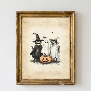 May include: A framed illustration featuring two frogs dressed in witch costumes and a ghost standing behind a carved orange jack-o'-lantern. The frogs wear pointed hats and capes, while the ghost is draped in a white sheet. Small bats fly above.