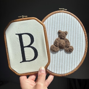 May include: Two embroidery hoops with wooden frames. One hoop is octagonal with a cream fabric background and a large black embroidered letter B. The other hoop is oval with a beige and white striped fabric background and a brown plush teddy bear.