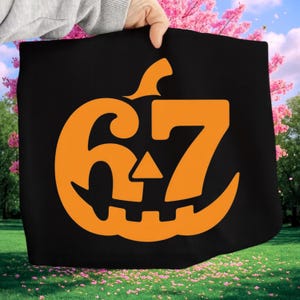 Pode incluir: Saco tote preto com um design de abóbora laranja e o número 67. A abóbora tem um rosto esculpido com um nariz triangular e um caule. O saco é exibido em um fundo de flores cor-de-rosa e grama verde.