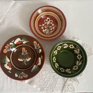 Puede incluir: Tres cuencos de cerámica decorativos con diseños florales. Uno es rojo oscuro con detalles amarillos y blancos, otro es marrón con flores naranjas y blancas, y el tercero es verde con flores blancas. Los cuencos están sobre un mantel de encaje blanco.