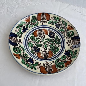 May include: A round, hand-painted ceramic plate with a cream base. It features a floral design with brown, blue, green, and orange accents. The rim has a blue border, and the style is rustic and decorative. The plate is approximately 25 cm in diameter.