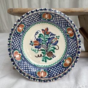 May include: A round ceramic plate with a cream-colored base featuring a hand-painted floral motif in blue, green, and orange. The rim displays a blue cross-hatched lattice pattern interspersed with orange and green circular floral accents.