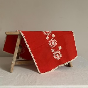 Può includere: Un runner da tavolo rosso ricamato con motivi floreali bianchi e un bordo all'uncinetto color crema. Il runner è drappeggiato su un piccolo sgabello di legno, mettendo in risalto i dettagli del ricamo.