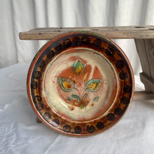 Op de afbeelding: Een handbeschilderde keramische plaat met een bloemenontwerp. De plaat heeft een crèmekleurig midden met een oranje en groen bloemmotief. De rand is bruin met donkerbruine stippen. De plaat wordt gepresenteerd op een houten oppervlak.