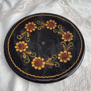 Puede incluir: Un plato decorativo redondo y negro con un diseño floral pintado a mano. Las flores son amarillas y rojas, con hojas verdes, dispuestas en un patrón circular. El plato tiene un borde dorado.