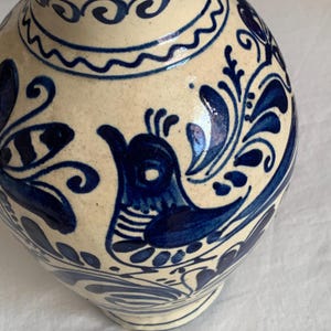 Pode incluir: Um vaso de cerâmica creme com intrincados desenhos florais e de pássaros azuis. O vaso apresenta um corpo arredondado e um pescoço estreito, mostrando a estética da arte popular.