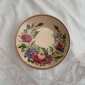 May include: A small, round, cream-colored plate with a hand-painted floral design. The flowers are in shades of pink, purple, blue, and yellow, with green leaves. The rim is a dark brown color. The plate is likely vintage or antique.