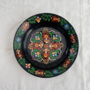 Könnte beinhalten: Ein schwarzer dekorativer Teller mit handgemaltem Blumendesign. Der Teller hat ein zentrales Blumenmuster in Rot, Orange, Gelb und Weiß, umgeben von einem Rand aus grünen Blättern und bunten Blumen. Der Teller ist rund.