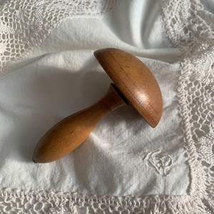 May include: A vintage wooden mushroom-shaped object with a handle. The mushroom top is rounded and the handle is cylindrical. The object is a light brown color and appears to be made of wood. It rests on a white cloth with lace trim.