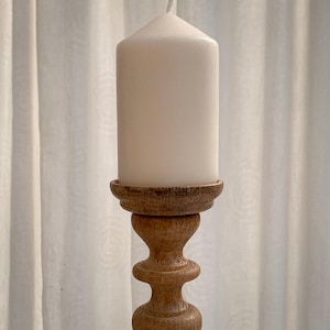 May include: A white pillar candle on a decorative wooden candlestick. The candlestick features a turned design with a round base and a detailed, textured top. The candle is unscented and cylindrical. The candlestick is made of wood.