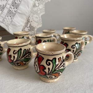 Può includere: Una collezione di piccole tazze in ceramica color crema con manici. Ogni tazza è decorata con un motivo floreale dipinto a mano in rosso, verde e arancione. Le tazze sono disposte su una superficie bianca, suggerendo un'estetica rustica o vintage.