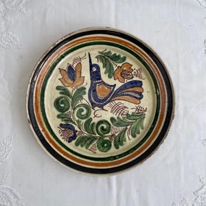 May include: Round ceramic plate with a hand-painted design featuring a blue bird, orange and blue flowers, and green leaves. The rim has black, green, and orange bands. The plate is on a white surface.