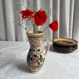 May include: A decorative, hand-painted ceramic pitcher with a handle, filled with red poppies and white baby's breath. The pitcher has a cream base with brown, blue, and green floral patterns. A stack of colorful ceramic plates is to the right.