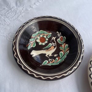 May include: A small, round, brown ceramic bowl with a hand-painted bird and floral design. The bird is white with orange and green accents. The rim has a wavy white and brown pattern. The bowl is on a white surface.