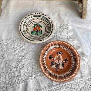 以下が含まれることがあります： 手描きのデザインが施された2つの丸い陶器のボウル。上のボウルはクリーム色で、緑とオレンジの花のモチーフと黒い線のディテールが特徴です。下のボウルはテラコッタ色で、黒と白の花柄が描かれています。