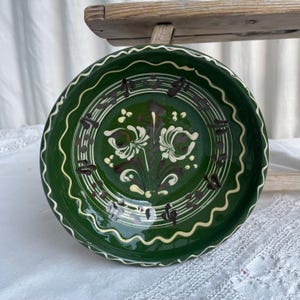 Op de afbeelding: Een ronde keramische kom met een diepgroen glazuur en een handgeschilderd bloemmotief in crème en donkerbruin. De binnenrand is versierd met een golvende crèmekleurige lijn, terwijl het midden een symmetrisch bloemmotief met bloemblaadjes toont.