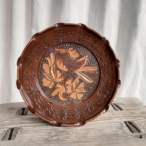 May include: A round, wooden decorative plate with a carved bird and floral design. The plate is brown with a scalloped edge and intricate detailing. The bird and flowers are lighter in colour, creating a contrast.