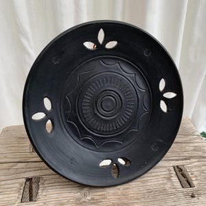 May include: A black decorative plate with a central circular design and cut-out floral shapes around the rim. The plate has a matte finish and sits on a weathered wooden surface. The plate is likely used for decorative purposes.