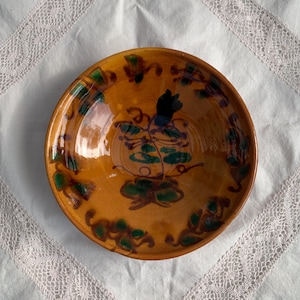 Op de afbeelding: Een ronde, geglazuurde keramische kom met een pompoenontwerp. De kom heeft een warme, oranje-bruine basiskleur met groene en blauwe accenten in het pompoen- en bladerontwerp. De rand van de kom heeft een decoratief patroon in groen en bruin.