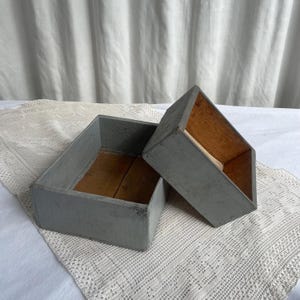 Könnte beinhalten: Zwei rechteckige Holzkisten mit grau lackierter Außenseite und naturbelassenem braunem Holz im Inneren. Die größere Kiste steht horizontal, während die kleinere Kiste schräg an der Seite des größeren Behälters angelehnt ist.