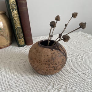 Pode incluir: Um pequeno vaso redondo de cerâmica marrom com interior e detalhes externos marrom escuro. O vaso contém vários caules de flores marrons secas. O vaso está sobre uma toalha de mesa branca rendada. Decoração de casa.