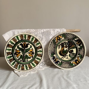 May include: Two decorative ceramic plates with hand-painted designs. One plate features a floral motif with yellow and black flowers, while the other showcases a bird design. Both plates have a cream-coloured background with green and brown accents.