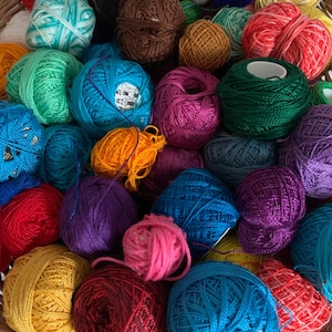 May include: A close-up of a basket filled with balls of yarn in a variety of colours, including blue, green, red, purple, yellow, and pink. The yarn is tightly wound, suitable for knitting or crochet.