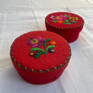 Op de afbeelding: Twee ronde rode vilten doosjes met deksels, elk voorzien van een handgeborduurd bloemmotief in paars, oranje, geel en groen. De randen van de deksels zijn versierd met een groen en geel zigzagsteekpatroon rondom de gehele omtrek.