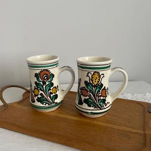 May include: Two ceramic mugs with cream-colored bodies and floral designs in orange, yellow, and green. Each mug has a handle and a green rim. The mugs are displayed on a wooden tray.