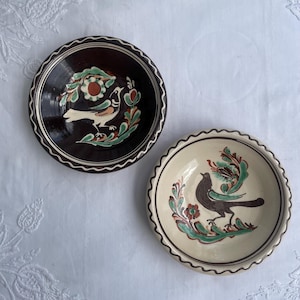 May include: Two ceramic bowls with hand-painted bird and floral designs. One bowl is dark brown with a white bird, the other is cream-colored with a dark bird. Both have scalloped edges and green, red, and white accents.