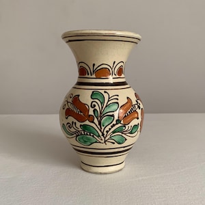 May include: A small, cream-colored ceramic vase with a flared top and base. The vase is adorned with hand-painted floral designs in shades of orange, green, and brown. The vase has a bulbous body and is decorated with horizontal brown stripes.