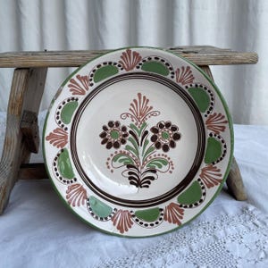 Op de afbeelding: Een ronde keramische bord met een crèmekleurige basis en een centraal handgeschilderd bloemmotief in bruin, groen en gedempt rood. De rand is versierd met afwisselende groene halve cirkels en rode waaiervormige patronen met bruine accenten.