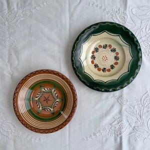 May include: Two round ceramic plates with decorative patterns. One plate features a terracotta base with green and white concentric rings and a central star motif. The other plate has a cream center with floral accents and a wide dark green rim.