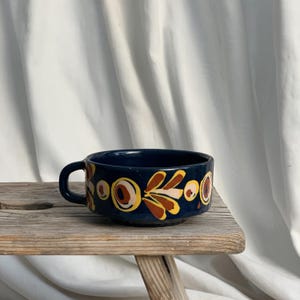 Può includere: Una ciotola in ceramica blu navy con manico, decorata con un motivo ripetuto di motivi floreali marroni, gialli e rosa. La ciotola è appoggiata su uno sgabello di legno rustico su sfondo bianco.