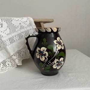 Pode incluir: Um jarro de cerâmica castanho escuro com uma pega, decorado com desenhos florais brancos e azuis e folhas verdes. A borda é branca com detalhes castanhos. O jarro tem um aspeto artesanal, rústico e vintage.