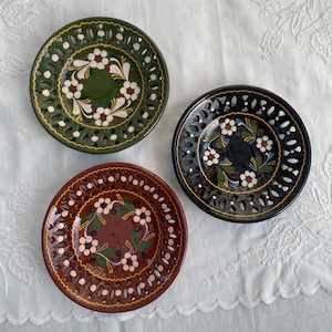 May include: Three decorative ceramic plates with floral designs. One plate is dark green, another is dark brown, and the third is black. Each plate features a pierced rim and a central floral motif in white and other colours.