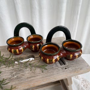 May include: Two ceramic planters with a handle, each holding two small pots. The pots are reddish-brown with yellow and green decorative patterns. The handles are black. The planters are on a rustic wooden surface.