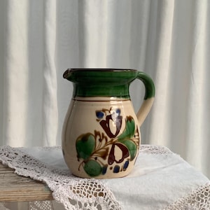 May include: A ceramic pitcher with a cream-colored body, a green rim and handle, and a spout. The pitcher is decorated with a floral pattern in shades of green, brown, and blue. The pitcher is sitting on a white lace doily.