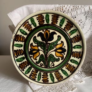 May include: A round, ceramic plate with a cream-colored background and a detailed floral design. The central motif features yellow and blue flowers with green leaves. The border has green and brown decorative elements. The plate is likely vintage.