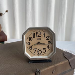 May include: Vintage octagonal alarm clock with a beige face, black numerals, and silver-colored metal frame. The clock face has the word "MOM" printed on it. The clock is set on a brown trunk.