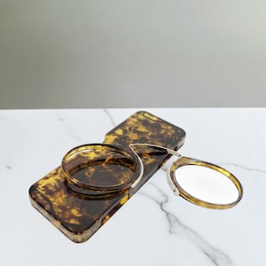 Puede incluir: Un espejo compacto y estuche con estampado de carey. El espejo ovalado tiene un marco plateado. El estuche rectangular y el espejo comparten un diseño de carey marrón y amarillo. El espejo está abierto, mostrando la superficie reflectante.