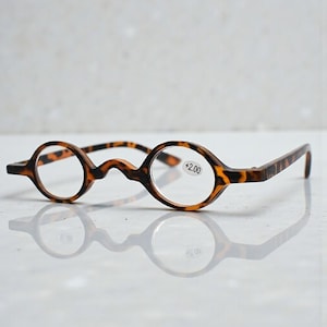 Puede incluir: Gafas de lectura redondas, con montura de carey y graduación +2.00. Las gafas tienen un diseño de puente único y varillas rectas. Las monturas son marrones con motas negras.