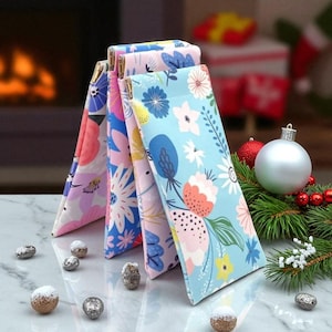 Puede incluir: Cuatro estuches para gafas con estampado floral en rosa, azul y amarillo. Los estuches tienen detalles dorados. En el fondo, decoraciones navideñas, como bolas y vegetación, sobre una superficie de mármol.