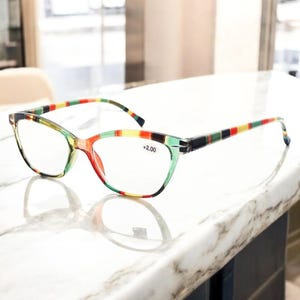 May include: Cat-eye reading glasses with a colourful striped pattern on the temples. The frames are a translucent green and red, with a +2.00 magnification. The glasses sit on a white marble surface.